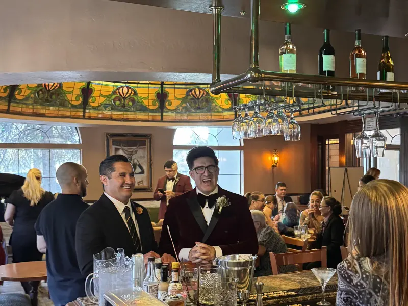 Two men in formal wear, one in a burgundy velvet dinner jacket, laughing at the bar beneath the stained-glass dome